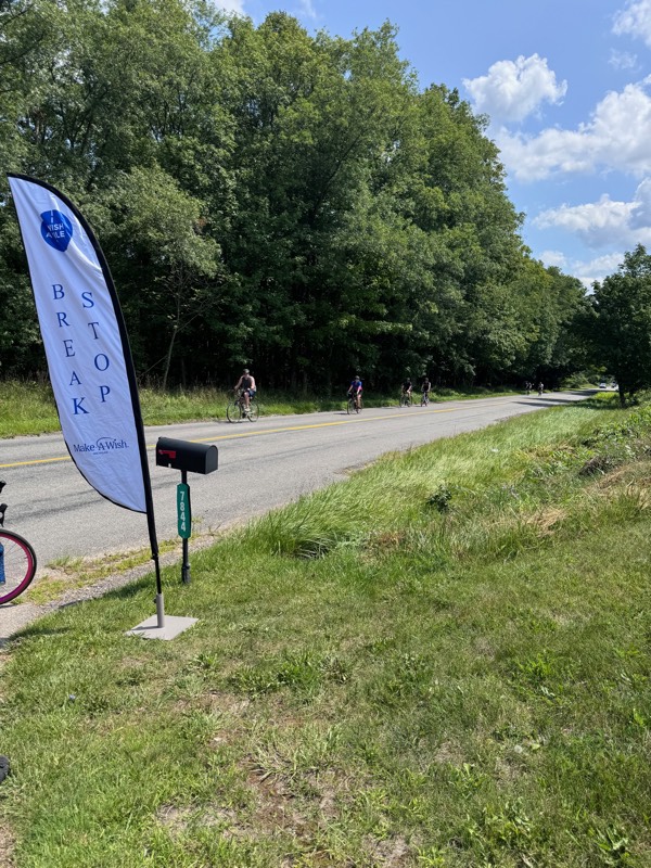 A Make-A-Wish "Break Stop" flag on a sunny country roadside as cyclists ride past.