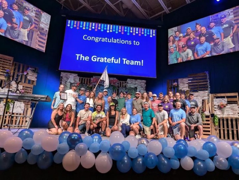 The Grateful Team gathered on a balloon-decorated stage in front of a screen reading "Congratulations to The Grateful Team!"