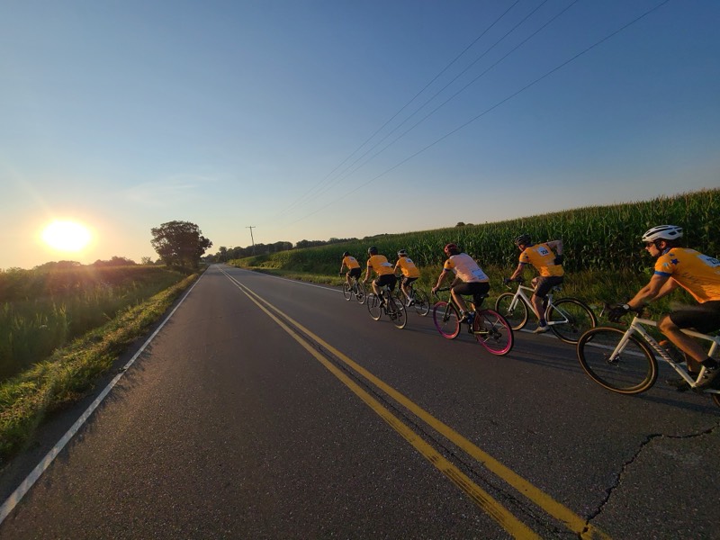 A line of riders silhouetted against a sunrise on a country road, golden light spilling across cornfields.