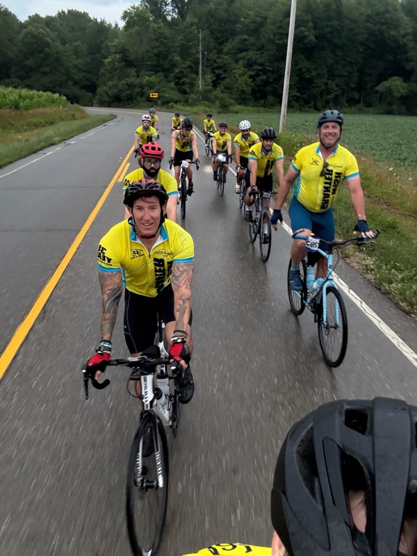 A group of WAM riders in yellow jerseys riding in formation on a wooded country road.