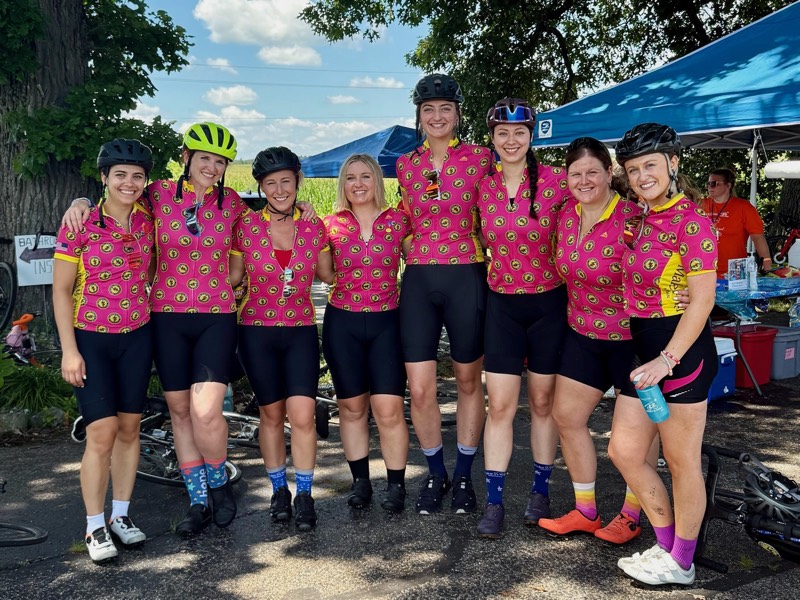 Eight Grateful Team riders in pink and yellow jerseys posing together at a rest stop.